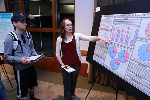 Female motioning to information on her poster, explaining it to an interested student.