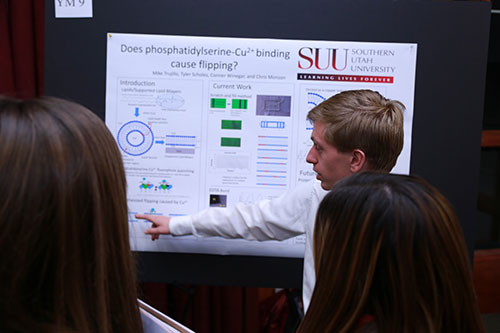 Male student presenting information on his poster to two other students.