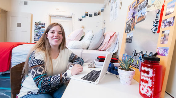 Student in dorm room, sitting at desk with notebook, facing the camera and smiling