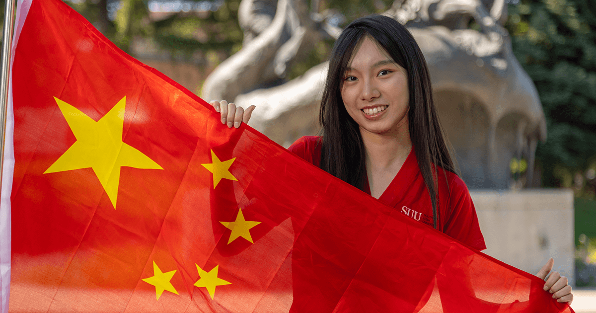 An international student smiles while holding the flag of their home country.