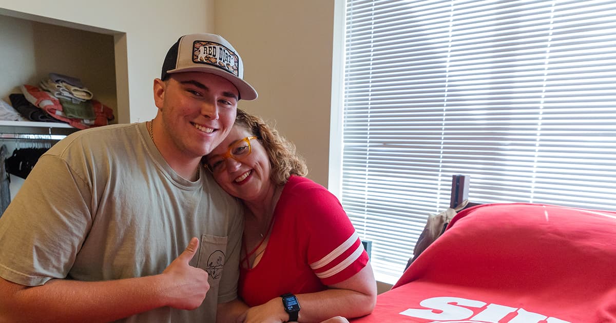 SUU student gives a thumbs up to the camera while embracing his mother in a college dorm room.