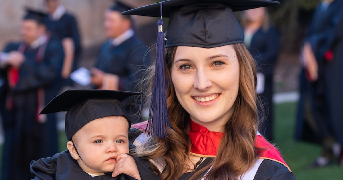 SUU student in graduation cap and gown holding her young child and smiling to the camera