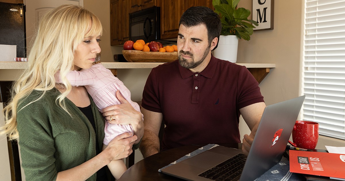 Two non-traditional students look at a laptop, while one holds and infant on her shoulder. 