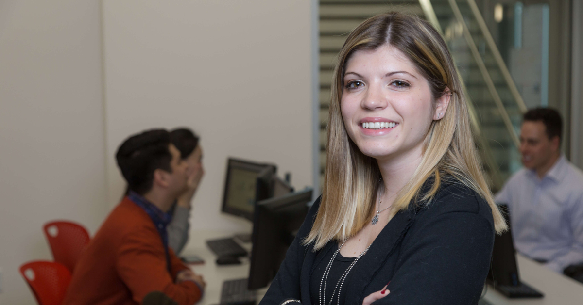 A student intern smiles into the camera with her coworkers in the background.