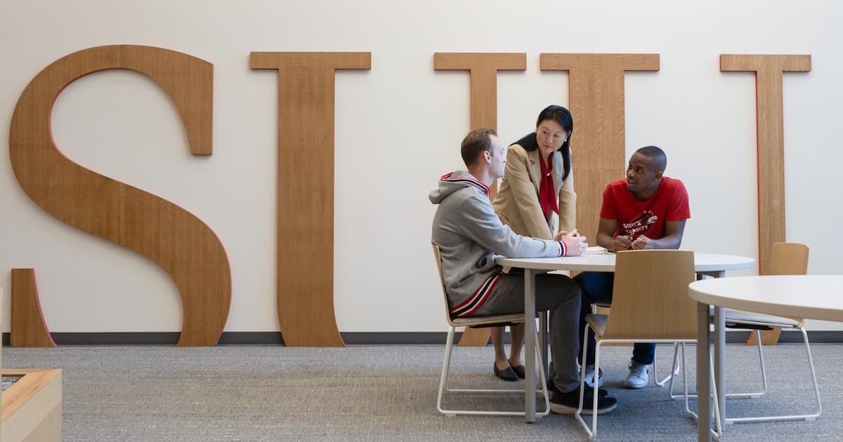 Students sitting at a table talking with a faculty member. They are on S.U.U. campus, indoors. On the wall behind them are large “S.U.U.” letters is natural colored wood. 
