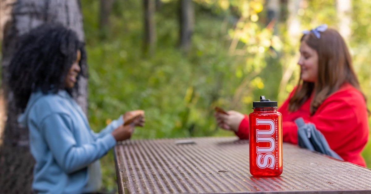 Two students sitting at a picnic table outside. On the table is a red water bottle with the letters “S.U.U.” on the side. 
