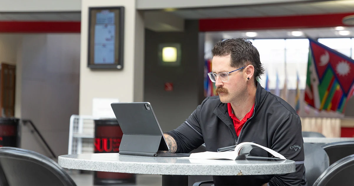 A student studies at a table with a tablet and a book.