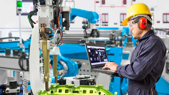 A man in a worker's jumpsuit and hard-hat holding a laptop in front of some complex machinery