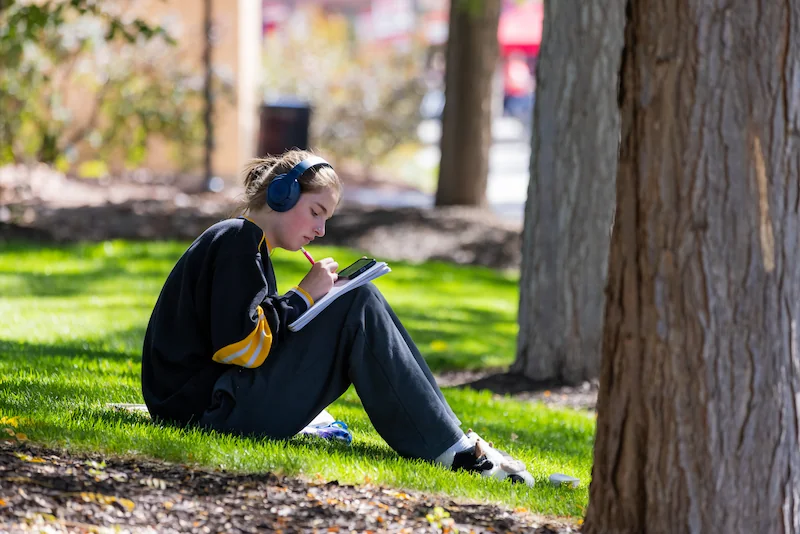 Student writing in a notebook and wearing headphones on the grass of SUU's upper quad between two trees