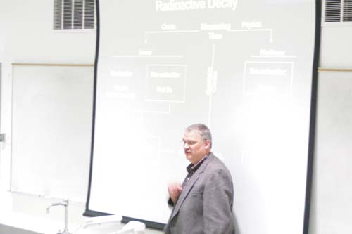 A man in front of a projector screen with a diagram on radioactive decay