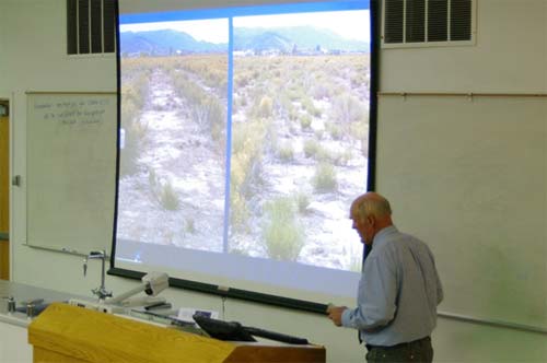An older man at the front of the classroom with two pictures of a desert