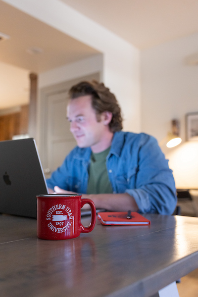 Male SUU student sitting at table with laptop and red coffee mug.