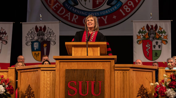 President Mindy Benson giving her inauguration address at the podium. She is wearing her inauguration regalia and the presidential seal of SUU is visible in the background.