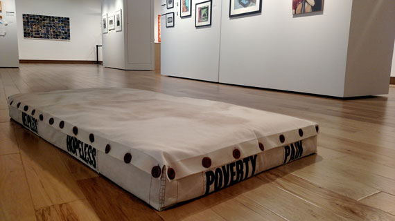 A mattress with the words helpless, hopeless, poverty, and pain on the sides. Artwork by Stuart Robinson.