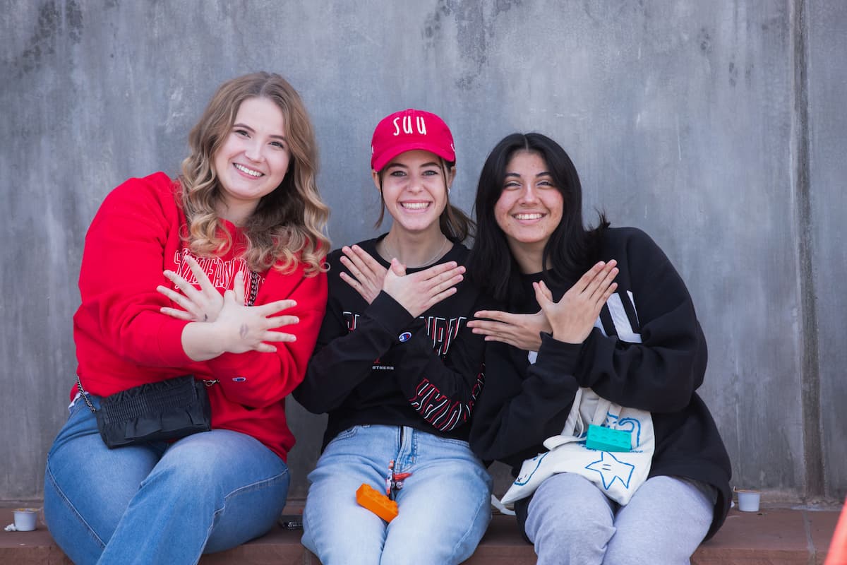 Three SUU students smile into the camera while making the T-Bird symbol with their hands. 