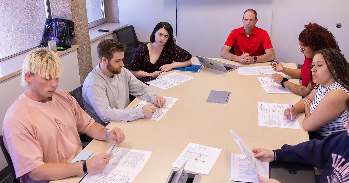 Students sit around a table studying papers with an instructor. 