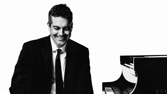 Black and White image. Jason Hardink looks down as he sits next to an open Steinway Grand Piano.