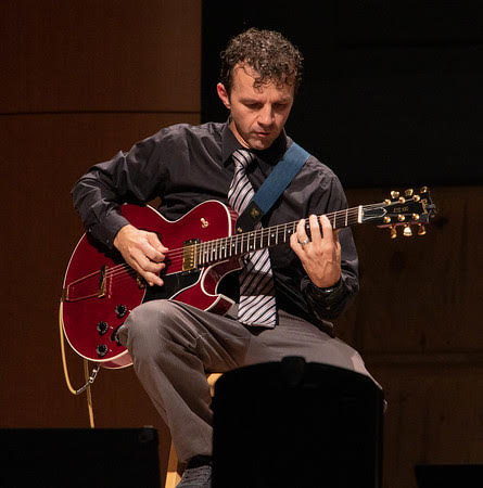 Dr. McKay Tebbs plays guitar while sitting on a stool.