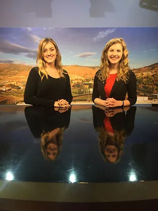 Two women sit behind a glossy news desk in a television studio