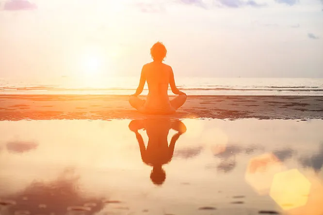 A woman is sitting on a beach in a meditative pose. Her reflection shines in a pool of water behind her. The sun is rising across the ocean in the background.