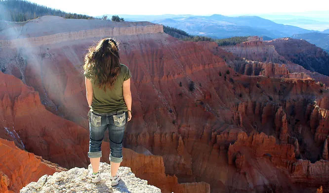 A view of a woman standing atop a cliff. There are red mountains in the background.