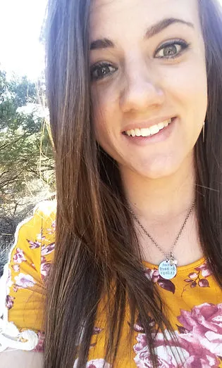 A selfie of a woman smiling. She is wearing a necklace and a yellow top with pink flowers on it.