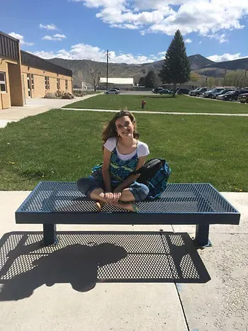 A young woman sits on a bench with her legs crossed. She is smiling while the wind blows through her hair. There is a large building to the left, and a mountain in the far distance