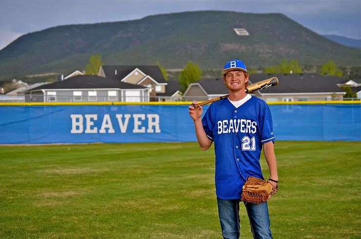 A man is posing in a baseball field. He is holding a baseball bat in his right hand, and a baseball glove in his left. There are houses in the distance, as well as a mountain with the letter “B” painted on it. A sign along the fence reads “BEAVER”