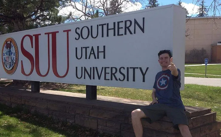 A young man smiling and giving the thumbs up while posing next to a sign that reads “SUU Southern Utah University”
