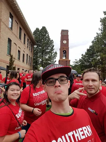 A photo of a young man making a silly face. There are crowds of people around him wearing red Southern Utah University shirts. The SUU clock tower is in the background.