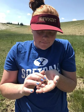 A person wearing a red “Viewmont” visor and a blue BYU softball T-shirt stands outdoors in a grassy, open landscape on a sunny day.
