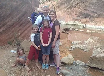 A family portrait featuring a man, wife, and four children. They are outside in a desert slot canyon. A small river flows past them.