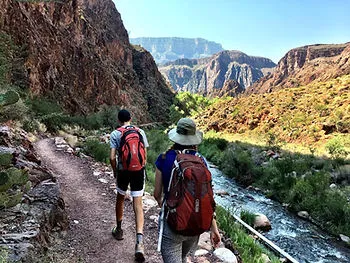 A man and a woman walk along a nature path. There is a river to the right of them, and a mountain on the left.