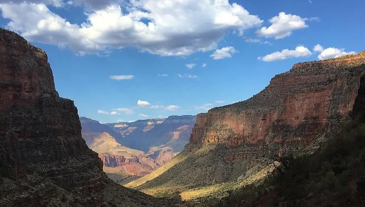 A wide view of a canyon landscape with steep, rugged rock walls on both sides and layered cliffs stretching into the distance. Sunlight illuminates parts of the valley floor, and a bright blue sky with scattered white clouds hangs overhead.