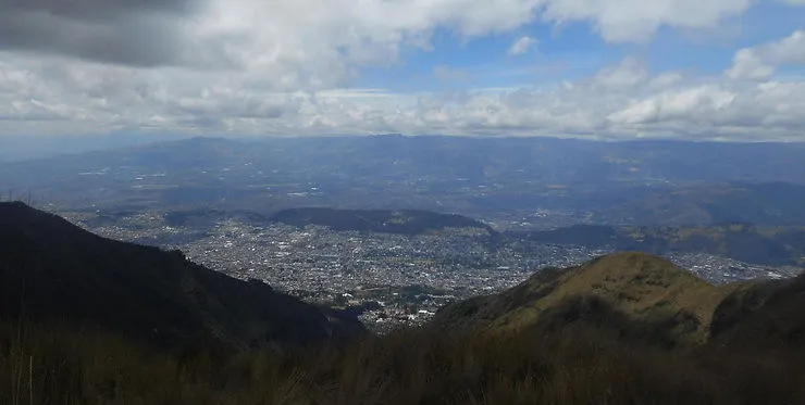 A high-altitude view overlooking a sprawling city nestled between mountain ridges. The foreground shows grassy slopes, while clouds cast shadows over the distant landscape under a partly cloudy sky.