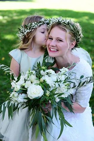 A smiling bride in a white dress holds a large bouquet of white flowers and greenery while a young girl wearing a flower crown kisses her on the cheek.