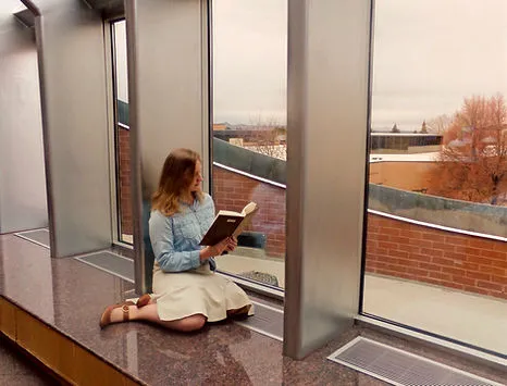 A person is kneeling on a bench next to a large window. They are reading a book.