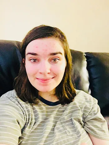 A person with shoulder-length brown hair sits on a dark leather couch, smiling softly at the camera. They wear a light gray and white striped shirt, and the background is a plain, softly lit wall.