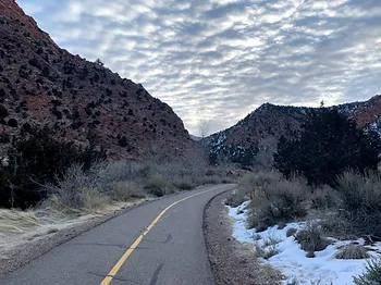 A paved walking trail extends into the far distance. Mountains are visible on both sides of the trail. Snow is covering the ground in patches.