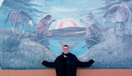A photo of a person posing in front of a traditional Native American painting. Their arms are outstretched in a gesture towards the painting