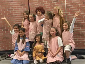 A group of children dressed in matching light pink costumes poses in front of a brick wall. They smile as they pose for a cast photo.