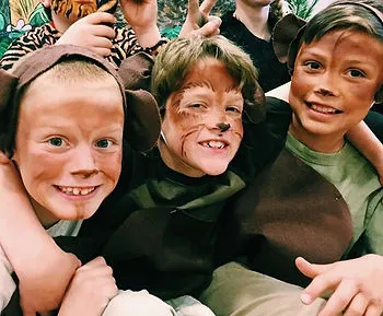 Three children dressed in brown costumes with face paint and animal ears pose together, smiling. They appear to be part of a theatrical production, sitting closely and looking at the camera.