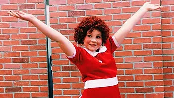 A child dressed as the character Annie stands in front of a brick wall, wearing a bright red dress with a white collar and belt. They smile widely with arms stretched high in a cheerful pose.