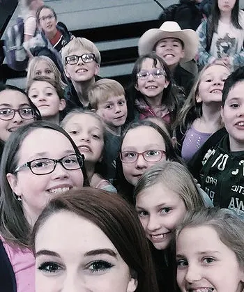 A group of children gathers closely for a selfie, smiling and looking at the camera. They stand indoors near bleachers, and the group includes kids of various ages, one of whom is wearing a cowboy hat.