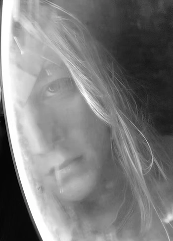 A black-and-white close-up photo of a person’s face reflected in or seen through a foggy, curved surface. Their long hair partially obscures one eye, and the lighting creates a tense, mysterious atmosphere.