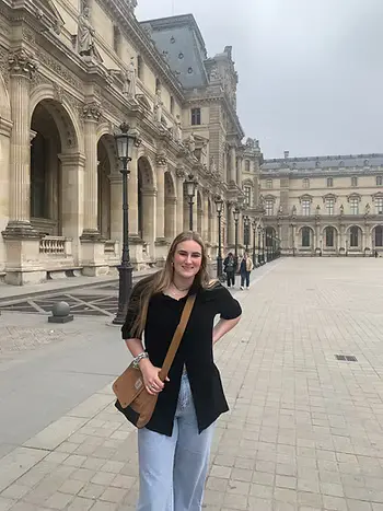 A woman stands smiling in a large stone courtyard with ornate historic buildings.