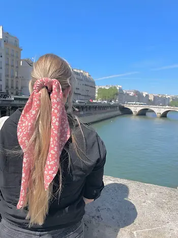 A close-up photograph taken from behind a young woman with long blonde hair tied in a high ponytail. A pink scarf with a small white floral pattern is tied around the ponytail, hanging down her back. She is standing on a stone bridge or railing overl