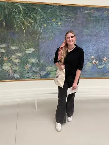 A young woman standing in an art museum in front of Claude Monet's large-scale water lily painting. She is smiling and wearing a black button-down shirt tied at the waist, wide-leg dark gray pants, and white sneakers. She has a pink floral-patterned