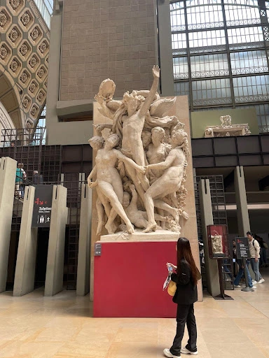 A woman stands before a tall marble sculpture of intertwined figures in a museum.