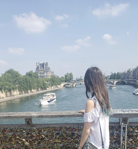 A woman looks out over a Paris river from a bridge covered in love locks. There are boats passing below in the river.